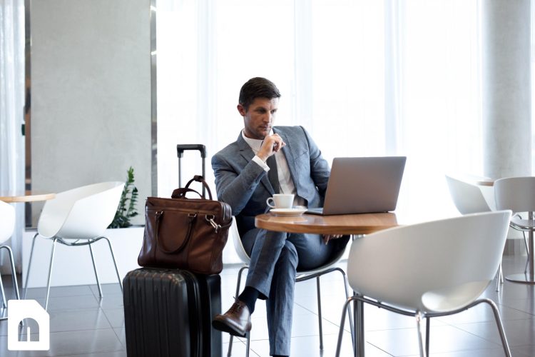 Zakenreiziger in een lounge, werkend op zijn laptop met een koffiekopje op tafel, terwijl zijn koffer en laptoptas naast hem staan.
